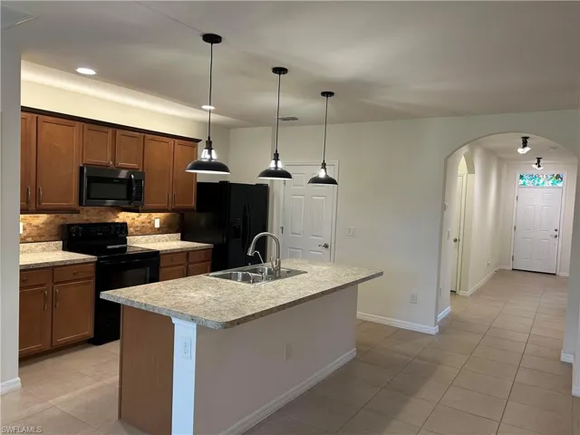 Kitchen featuring black appliances, backsplash, a center island with sink, arched walkways, and light tile patterned floors