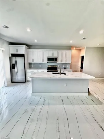 Kitchen featuring stainless steel appliances, white cabinets, light stone counters, a center island with sink, and recessed lighting