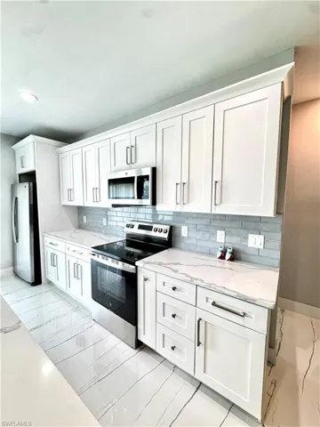 Kitchen featuring appliances with stainless steel finishes, light marble finish floors, backsplash, white cabinets, and light stone countertops