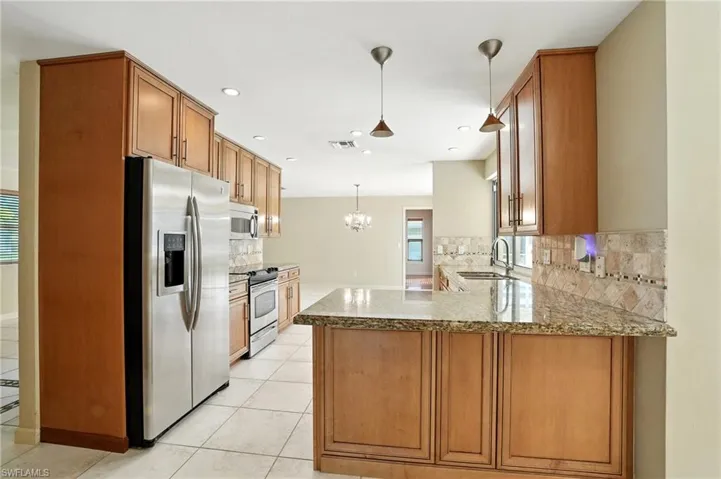 Kitchen with a peninsula, light stone countertops, stainless steel appliances, brown cabinetry, and decorative light fixtures