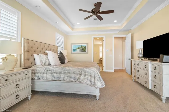 Carpeted bedroom featuring a tray ceiling, ceiling fan, crown molding, and ensuite bathroom