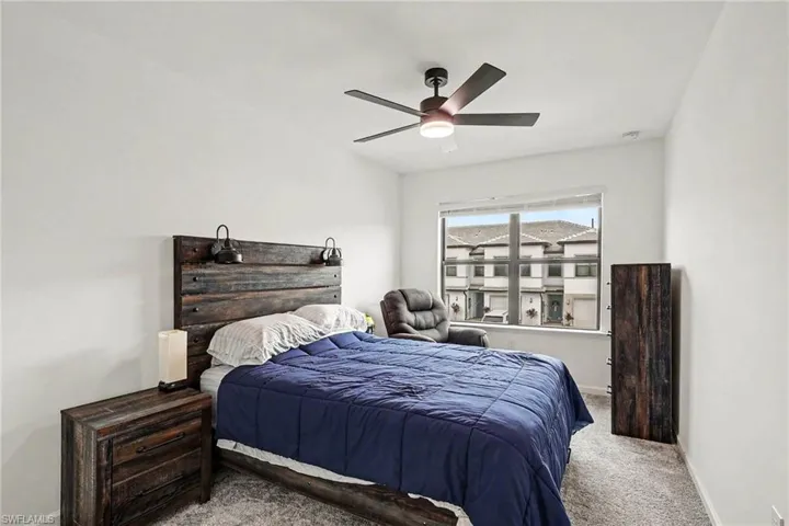 Bedroom featuring light grey carpeting, white painted walls, a large window, and a ceiling fan with integrated lighting