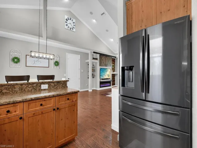 Kitchen with stainless steel fridge, dark stone counters, dark wood-style floors, brown cabinets, and hanging light fixtures