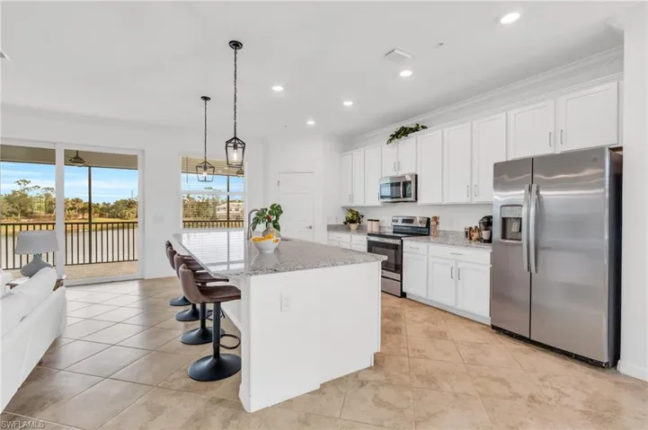 Kitchen featuring white cabinets, appliances with stainless steel finishes, decorative light fixtures, an island with sink, and light stone counters