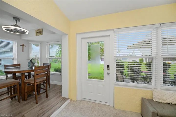 Wood-finish flooring in dining area, white framed windows with blinds, modern ceiling fan with integrated lighting, glass-paneled entry door, light-toned wall finishes