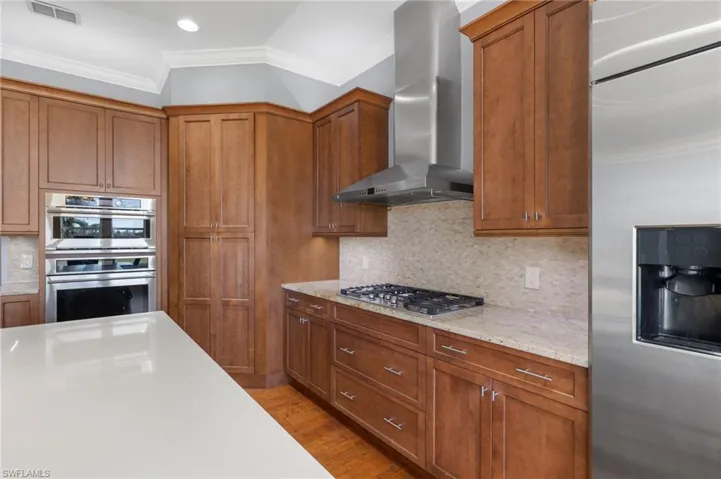 Kitchen with wall chimney range hood, ornamental molding, light wood-type flooring, appliances with stainless steel finishes, and tasteful backsplash
