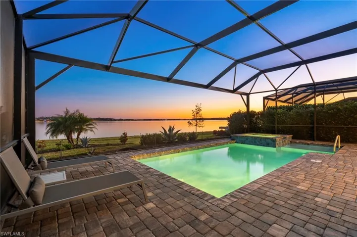 Pool at dusk with an in ground hot tub, a water view, glass enclosure, and a patio area