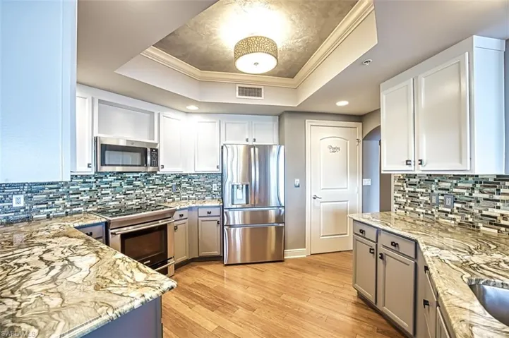 Kitchen with crown molding, stainless steel appliances, light wood finished floors, light stone countertops, and a raised ceiling