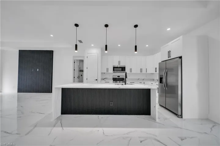 Kitchen featuring light stone countertops, appliances with stainless steel finishes, white cabinets, decorative light fixtures, and an island with sink