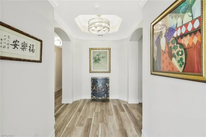 Corridor featuring ornamental molding, light wood finished floors, a chandelier, arched walkways, and a tray ceiling