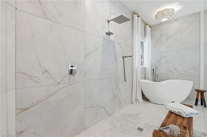Spacious bathroom featuring large format marble-look wall tiling, a rainfall shower head with handheld attachment, a freestanding soaking tub, and mosaic tile flooring