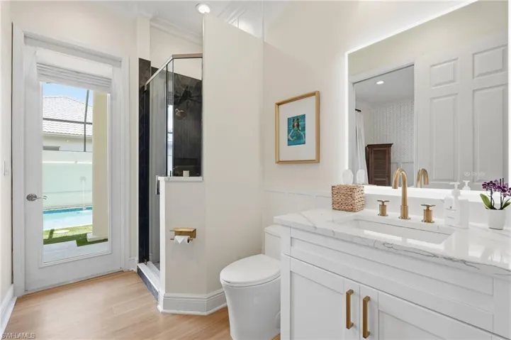 Bathroom featuring wood-finish flooring, a white single vanity with a stone countertop, gold-finish fixtures, and a frameless glass shower enclosure with dark tiling