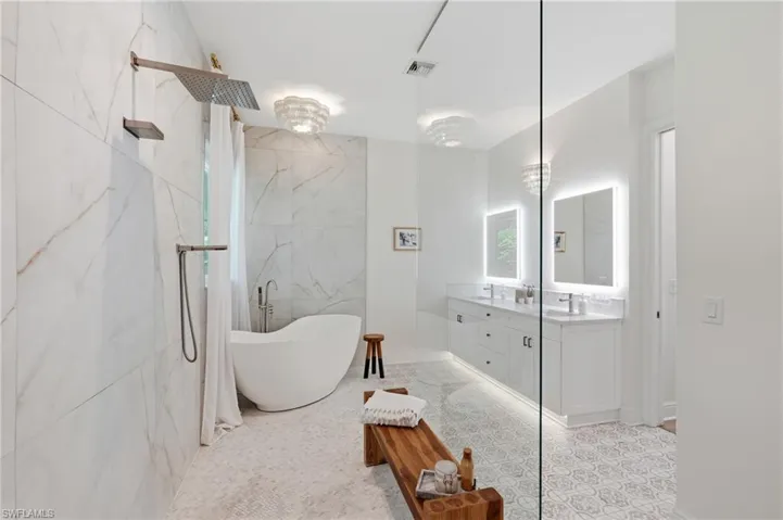 Spacious bathroom featuring a freestanding soaking tub, a rainfall showerhead, large format porcelain tile walls, a dual vanity with undermount sinks, and illuminated mirrors