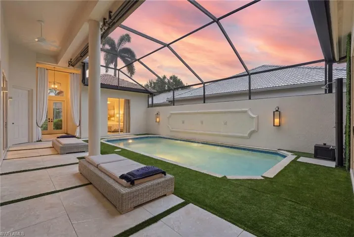 Screened-in outdoor pool area featuring a rectangular pool with a decorative wall fountain, light-toned large format tile patio, artificial turf accents, and an outdoor shower fixture
