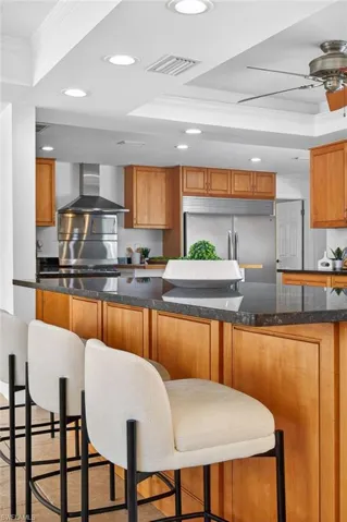 Kitchen with ornamental molding, a kitchen breakfast bar, a tray ceiling, ceiling fan, and dark stone countertops
