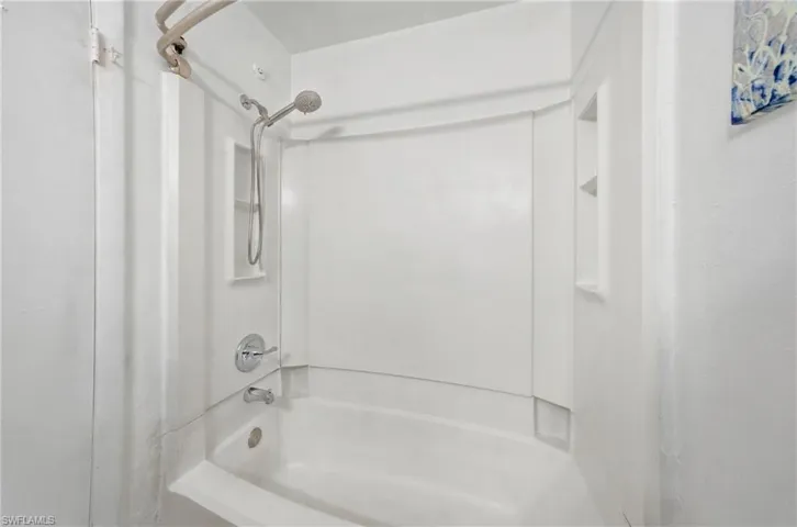 White acrylic tub and shower combination featuring a built-in shelf, brushed nickel shower head, and matching faucet with tub spout