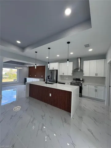 Kitchen with appliances with stainless steel finishes, decorative light fixtures, an island with sink, white cabinets, and wall chimney range hood