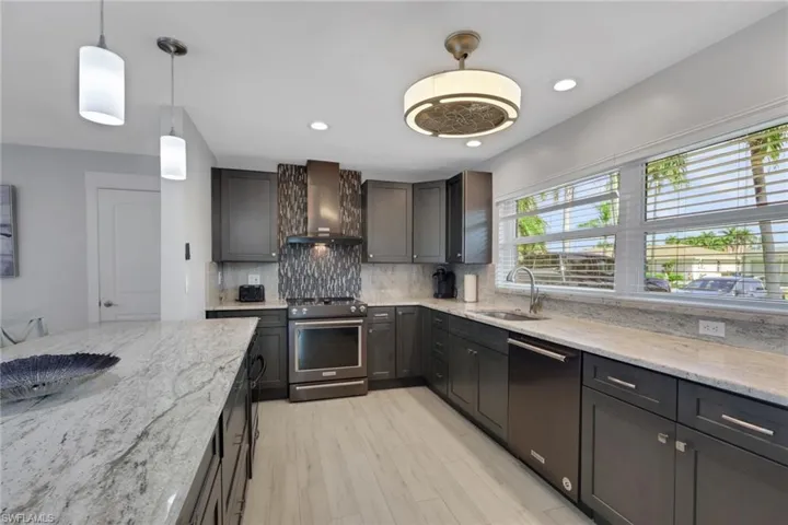 Kitchen featuring electric range, light stone counters, wall chimney range hood, dishwashing machine, and hanging light fixtures