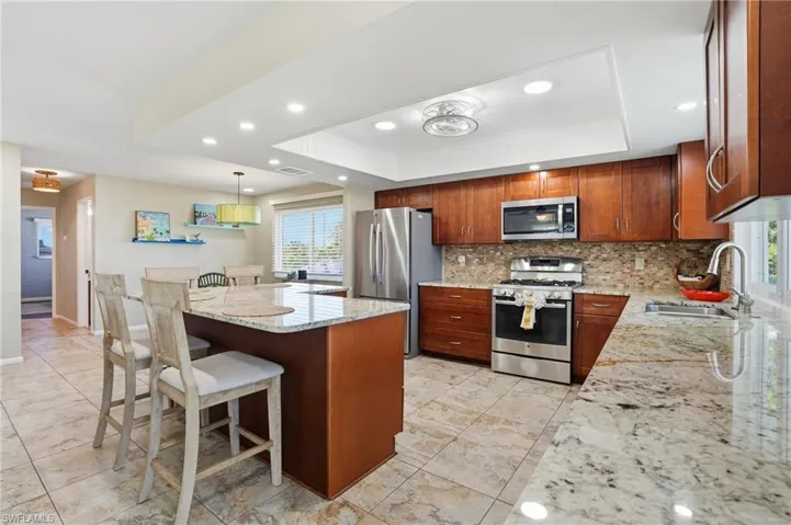 Kitchen with light stone countertops, stainless steel appliances, a kitchen bar, a raised ceiling, and recessed lighting - Virtually Edited Image
