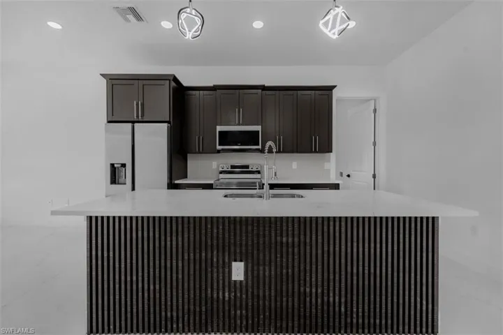 Kitchen featuring an island with sink, stainless steel appliances, light marble finish floors, light stone countertops, and dark wood finish cabinets