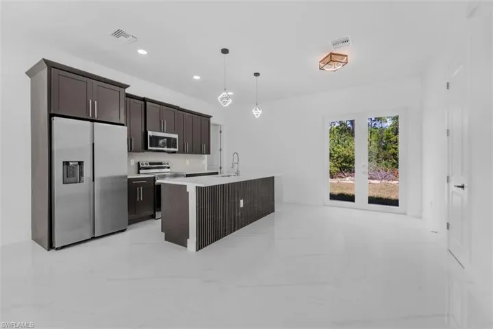 Kitchen featuring stainless steel appliances, dark wood finish cabinets, a center island with sink, decorative light fixtures, and light stone countertops