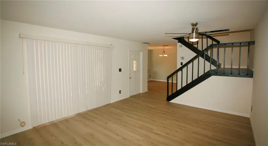 Living area featuring wood-finish flooring, vertical blinds on a large window, a ceiling fan with integrated lighting, and a prominent staircase with dark railings