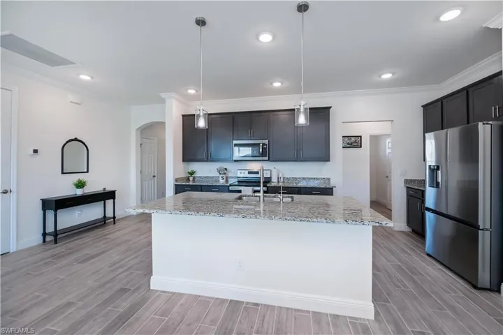 Kitchen featuring stainless steel appliances, crown molding, arched walkways, light stone counters, and an island with sink