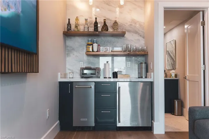 Indoor bar with open shelves, tasteful backsplash, stainless steel fridge, dark wood-style floors, and light stone countertops