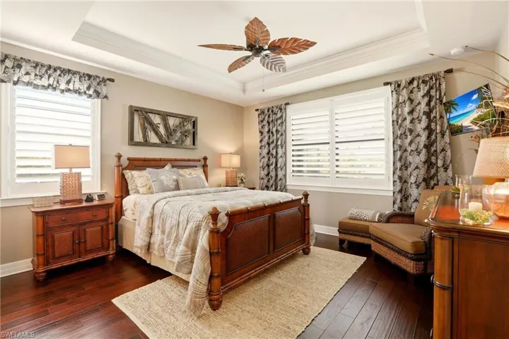 Primary Bedroom featuring dark wood laminate flooring, ornamental molding, multiple windows, a ceiling fan, and a tray ceiling