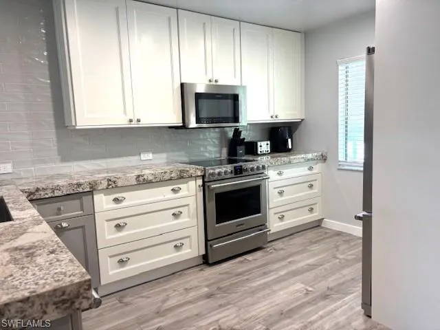 Kitchen featuring appliances with stainless steel finishes, white cabinetry, light stone counters, light wood finished floors, and backsplash
