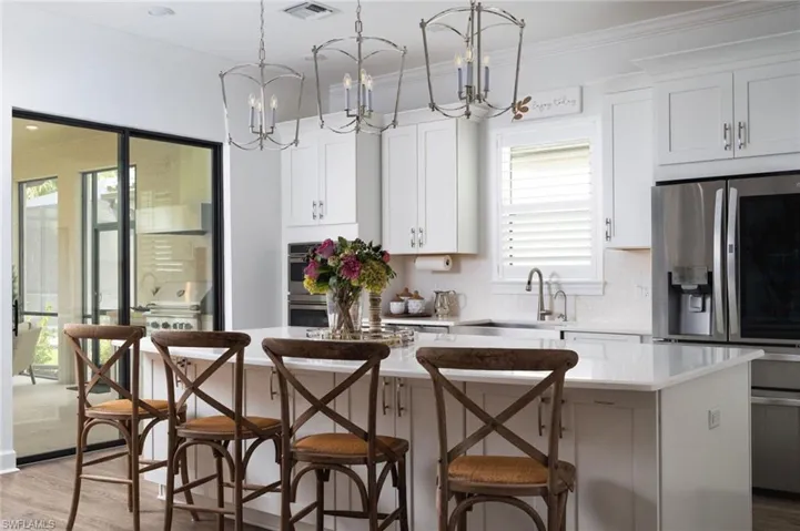 Bright kitchen featuring a white island with a light-toned countertop, white cabinetry with silver hardware, recessed lighting, and two ornate pendant light fixtures