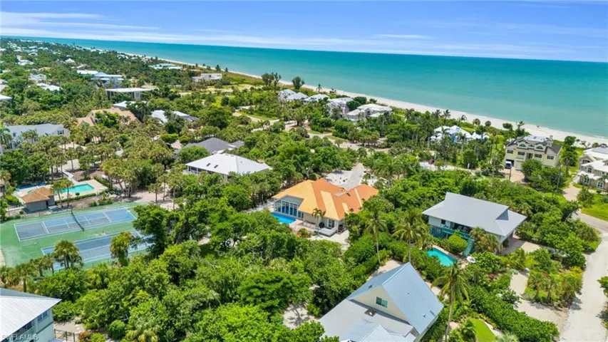 Aerial perspective showcasing a coastal community with numerous residences, two tennis courts, and a sandy beachfront bordering turquoise waters