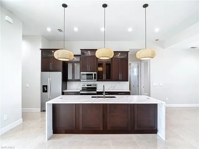 Kitchen featuring light stone countertops, an island with sink, pendant lighting, and appliances with stainless steel finishes