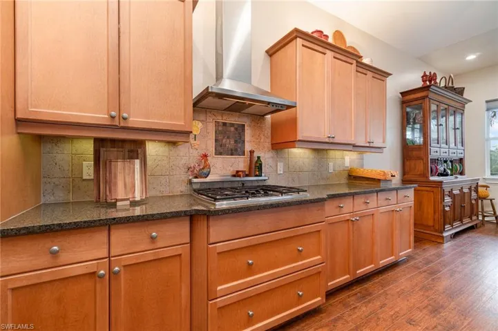 Kitchen featuring stainless steel gas stovetop, dark wood finished floors, dark stone countertops, wall chimney range hood, and backsplash