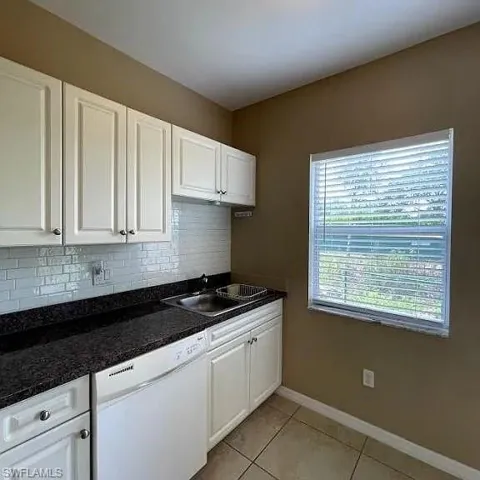 Kitchen featuring light tile patterned floors, white dishwasher, a sink, white cabinetry, and backsplash