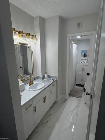White cabinetry with gold-tone hardware, solid-surface vanity countertop, undermount sink with gold-tone faucet, arched mirror, and stone-finish tile flooring