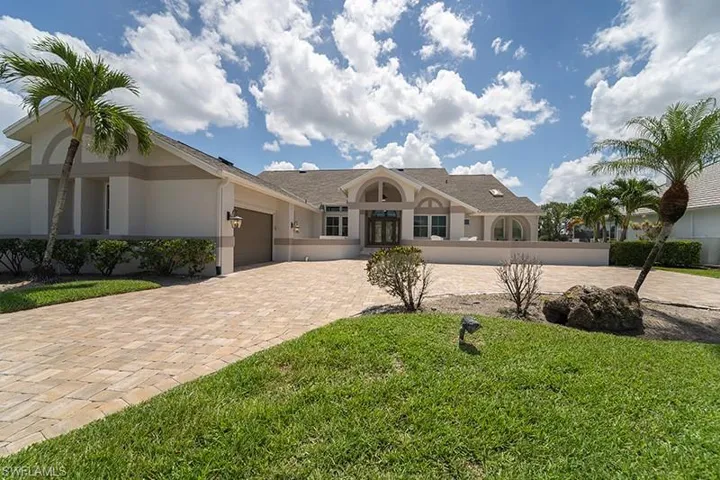 Expansive paver driveway leading to a residence featuring a stucco exterior with decorative banding, arched windows, and mature palm trees