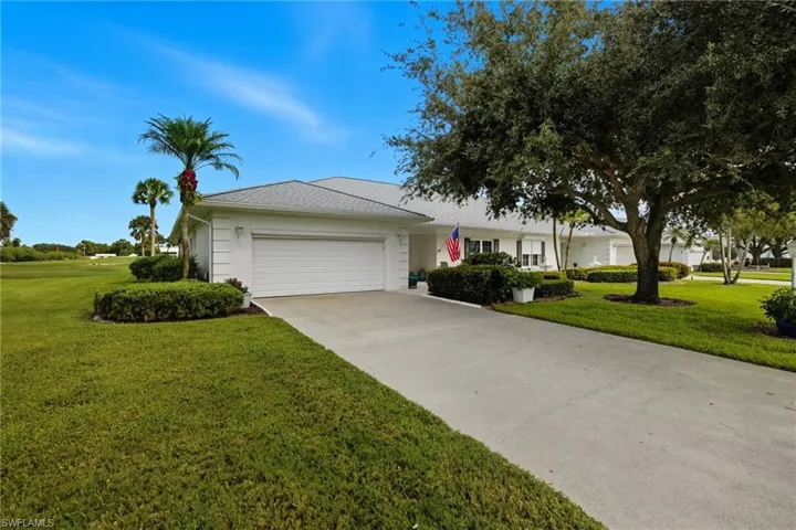 Single-story white exterior with a two-car garage, concrete driveway, and front lawn landscaping featuring mature trees and shrubbery
