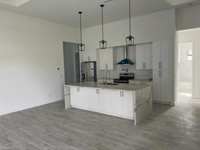 Kitchen with light stone counters, a large island with sink, wall chimney range hood, stainless steel appliances, and decorative light fixtures