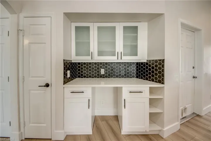Bar area with white cabinetry, light stone counters, light wood-style flooring, glass insert cabinets, and backsplash