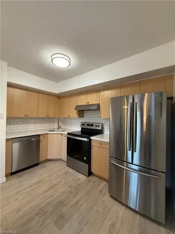 Kitchen with stainless steel appliances, a textured ceiling, under cabinet range hood, backsplash, and light brown cabinetry