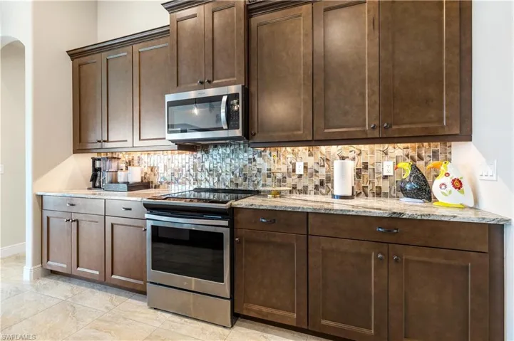 Kitchen featuring stainless steel appliances, decorative backsplash, light stone counters, dark brown cabinetry, and light tile patterned floors