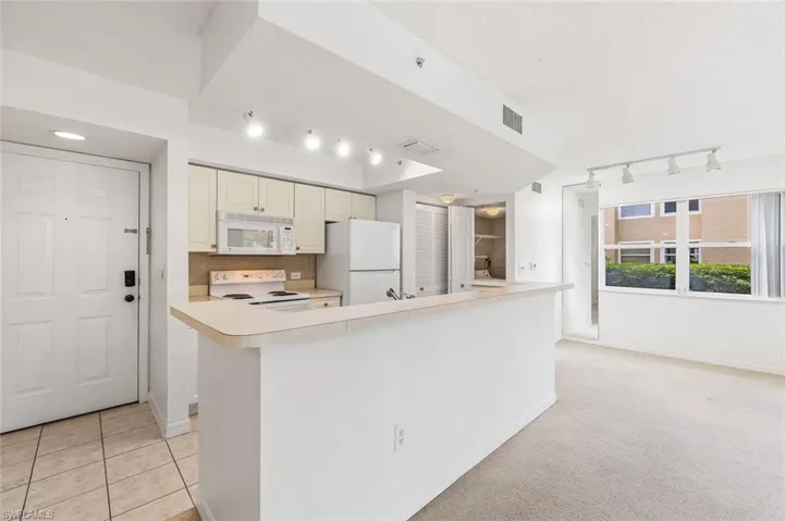 Open-concept living area featuring a kitchen with light-toned cabinetry, white appliances, and track lighting, and tile floors in the kitchen and carpet in the living areas.