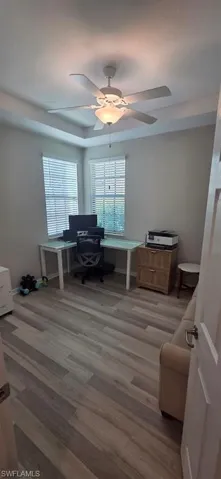 Office space with light wood-style flooring, ceiling fan, and a tray ceiling