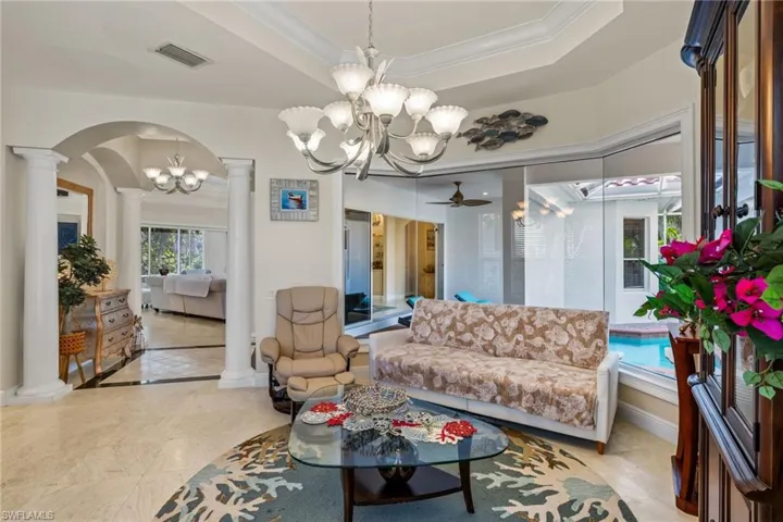 Living room with a raised ceiling, crown molding, a ceiling fan, ornate columns, and a chandelier