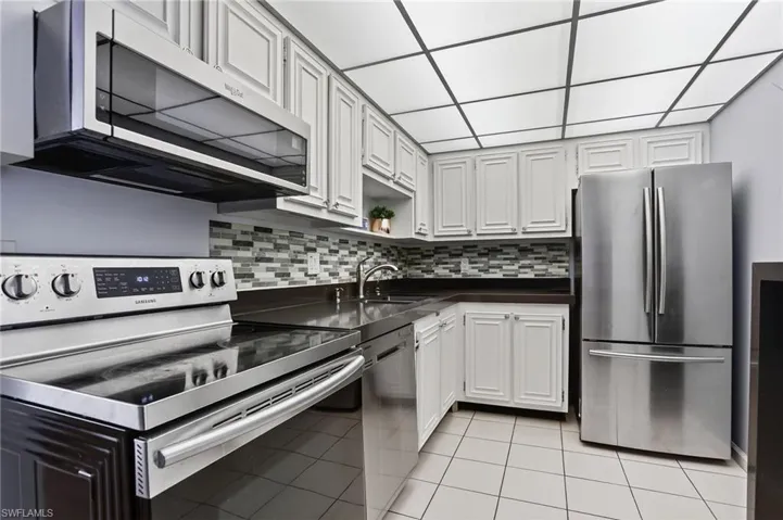 Kitchen featuring appliances with stainless steel finishes, light tile patterned flooring, dark countertops, backsplash, and white cabinetry