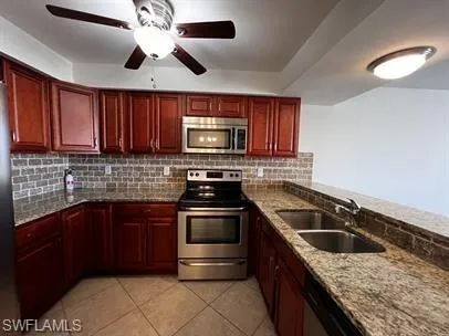Kitchen with stainless steel appliances, bold wood finish cabinetry, light stone countertops, ceiling fan, and light tile patterned flooring