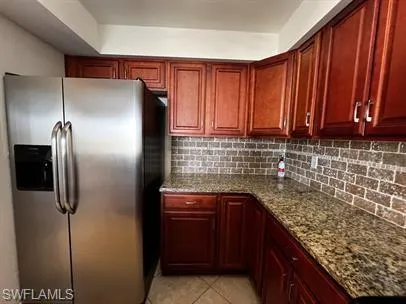 Kitchen featuring stainless steel fridge, bold wood finish cabinets, dark stone counters, light tile patterned floors, and decorative backsplash