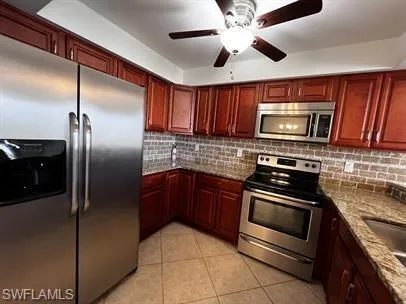 Kitchen featuring stainless steel appliances, light stone counters, bold wood finish cabinets, a ceiling fan, and light tile patterned floors
