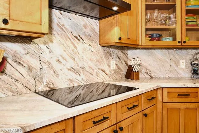 Kitchen featuring glass insert cabinets, black electric cooktop, light stone countertops, backsplash, and wood finish cabinets
